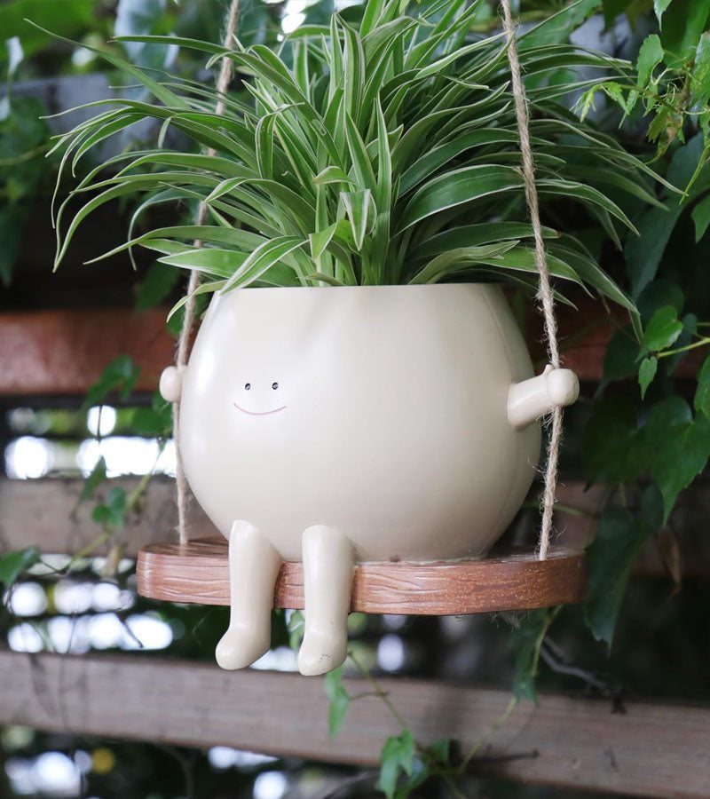 Cache-pot blanc souriant assis sur une balançoire en bois, avec une plante verte