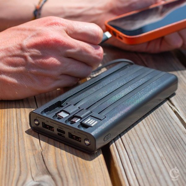 Batterie externe solaire noire avec câbles intégrés rechargeant un smartphone sur une table en bois