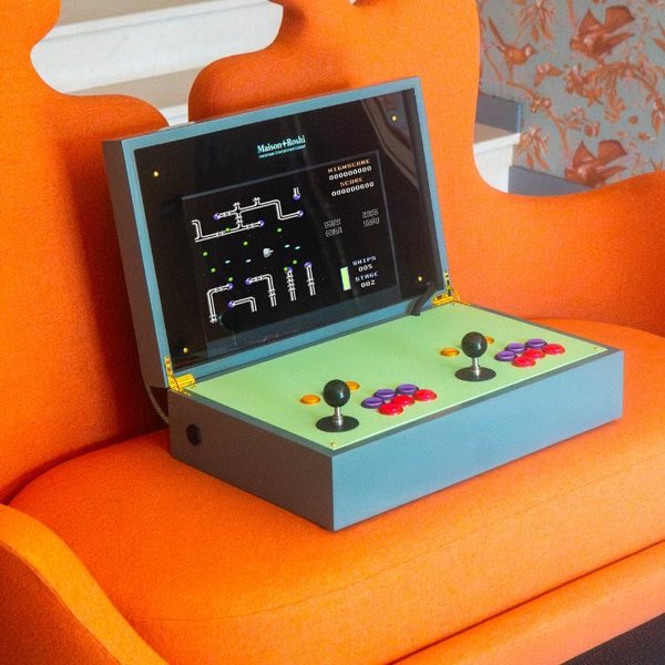 Borne d'arcade portable en bois avec écran ouvert et deux joysticks posée sur un fauteuil orange