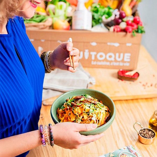 Femme tenant une assiette de nouilles aux légumes devant un carton Quitoque rempli d'ingrédients frais
