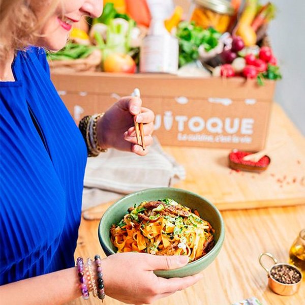 Femme tenant une assiette de nouilles aux légumes devant un carton Quitoque rempli d'ingrédients frais