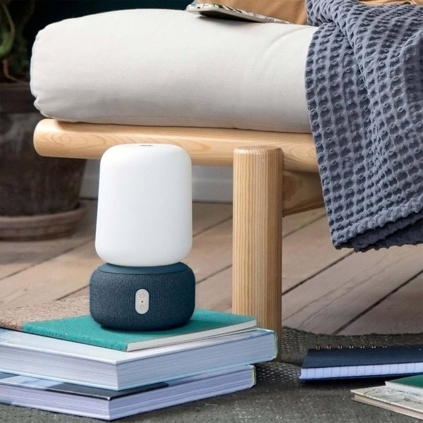 Enceinte lampe aLOOMI à abat-jour blanc et base noire posée sur une pile de livres près d'un fauteuil
