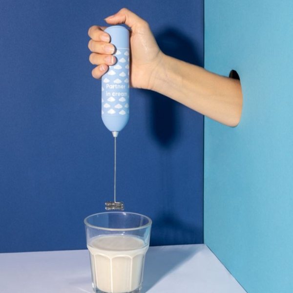 Main tenant un mousseur à lait bleu à motif nuages au-dessus d'un verre de lait mousseux
