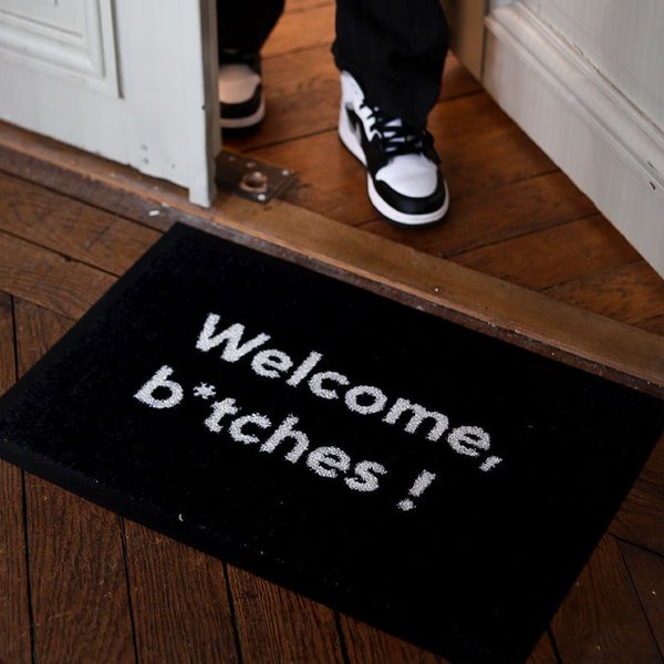 Paillasson noir « Welcome, b*tches ! » devant une porte d'entrée, avec un pied en baskets qui passe le seuil