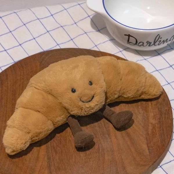 Peluche Jellycat en forme de croissant souriant avec petites jambes, posée sur une planche en bois