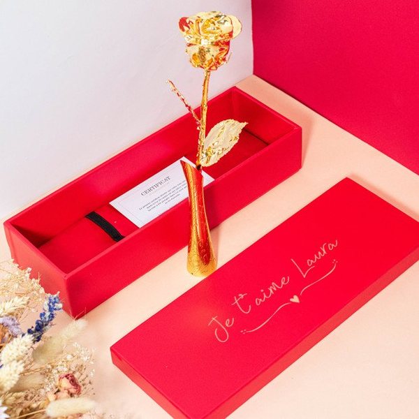 Rose éternelle dorée à l'or 24 carats dans son vase doré, posée devant un coffret rouge gravé « Je t'aime Laura »