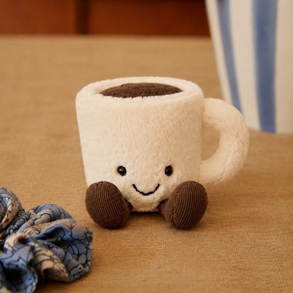 Peluche Jellycat en forme de tasse d'espresso souriante posée sur une table