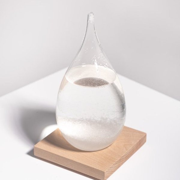 Storm glass en forme de goutte avec cristaux blancs posé sur un socle en bois clair
