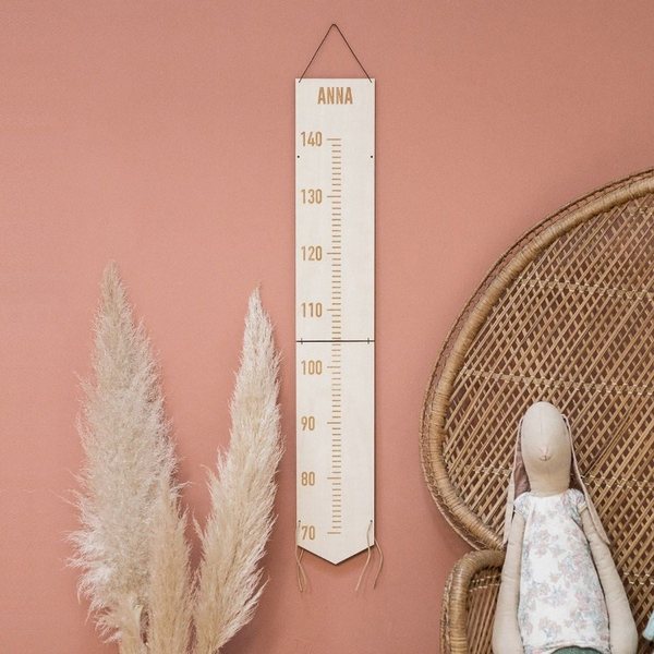 Toise en bois clair gravée d'un prénom accrochée sur un mur terracotta à côté d'un fauteuil en rotin