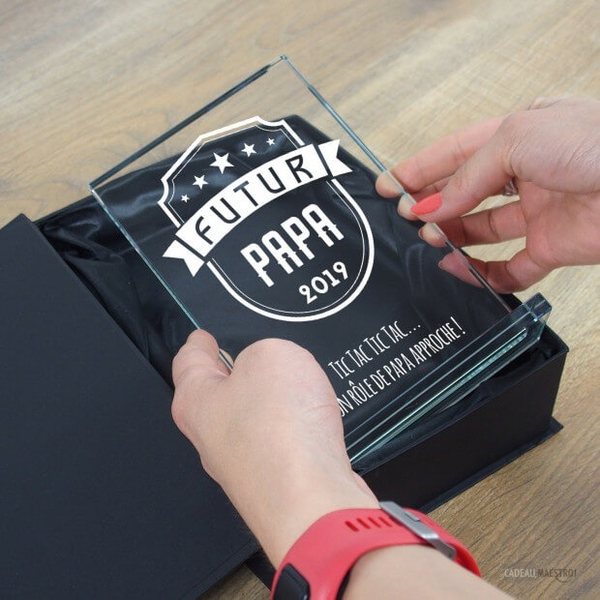 Mains sortant d'un coffret noir un trophée en verre gravé « Futur papa » avec l'année et une phrase