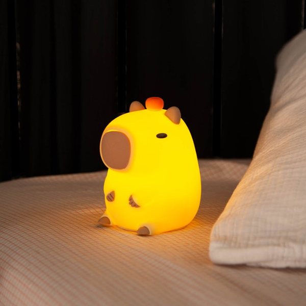 Veilleuse capybara jaune allumée posée sur un lit, lumière chaude dans une chambre sombre